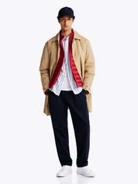 Beiger Übermantel über einer roten Puffweste, weiß-blau gestreiftes Hemd, marineblaue Hose und weiße Sneakers. Schwarze Mütze mit Logodetail.