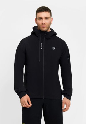 Mann trägt schwarze Kapuzenjacke mit Reißverschluss und schwarze Hose, steht vor schlichtem hellem Hintergrund.