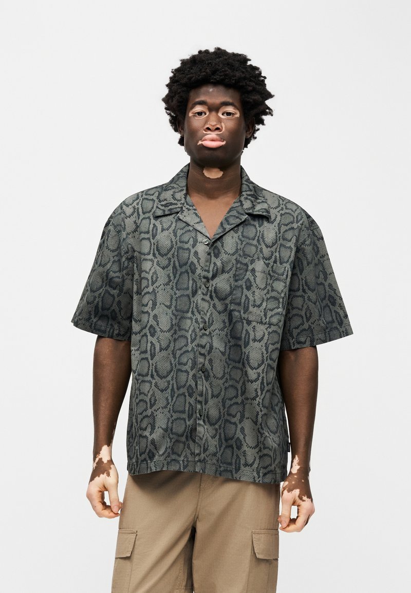 Camicia a maniche corte con stampa snake, caratterizzata da una combinazione di colori verde e nero, bottoni frontali e una tasca sul lato sinistro.