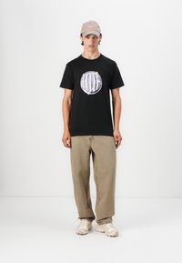 Mustast puuvillast T-särk, millel on ümmargune lillakas graafiline disain, koos beežide lahtiste pükste ja heledate tossudega. Peal beež nokamüts.