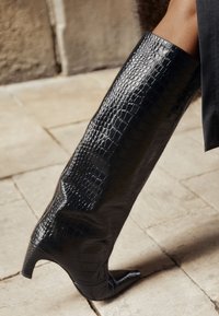 Crna čizma visoka do koljena, sa špicastim vrhom i niskom petom u obliku mačkica, teksture krokodilove kože, nošena na nozi koja korača po kamenoj površini.
