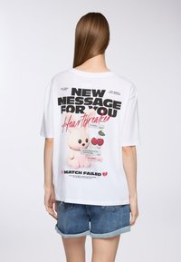 Biała, oversized koszulka z nadrukiem w postaci różowego króliczka, wiśni oraz czarnego i czerwonego tekstu, z postarzanym wykończeniem i luźnym krojem.