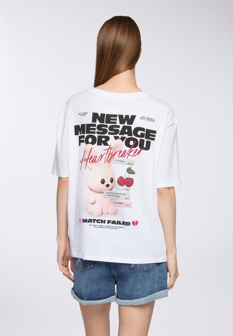Biała, oversized koszulka z nadrukiem w postaci różowego króliczka, wiśni oraz czarnego i czerwonego tekstu, z postarzanym wykończeniem i luźnym krojem.