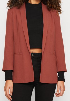 Femme portant un blazer ouvert couleur rouille sur un top court noir et un pantalon noir taille haute, avec des cheveux bouclés visibles aux épaules.