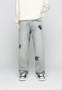 Persona che indossa jeans chiari con lettere ricamate nere, un felpa panna e sneakers bianco e nere, in piedi davanti a uno sfondo neutro.