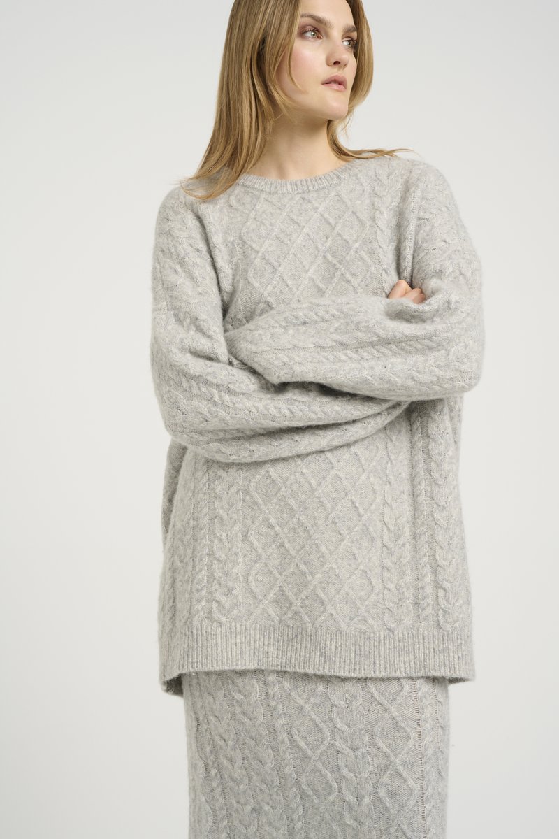 Set de pulover și fustă tricotate de culoare gri deschis, cu un model de tricot în carvăn. Puloverul are un decolteu rotund și un croi oversize. Textură moale și căldură.