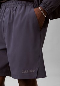 Άνδρας που φοράει σορτς Calvin Klein σκούρου γκρι με ελαστική μέση, χέρι στην τσέπη και ασημένιο βραχιόλι με αλυσίδα στον καρπό.