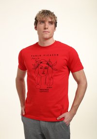Unge mand iført en rød Pablo Picasso t-shirt med en sort linjekunst tegning af et kvindeligt ansigt og grå bukser, stående med hænderne i lommerne.