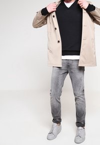 Trench coat beige sopra un maglione nero, jeans grigi e sneakers grigi. Il cappotto presenta bottoni grandi e una texture liscia.