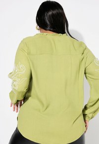 Camicia verde con bottoni, maniche lunghe, caratterizzata da ricami floreali su una manica e un colletto arricciato. Tessuto morbido e vestibilità comoda.
