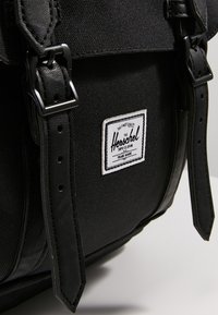 Czarny plecak Herschel z materiałowym korpusem, skórzanymi akcentami, regulowanymi paskami oraz białą łatą z logo z przodu.