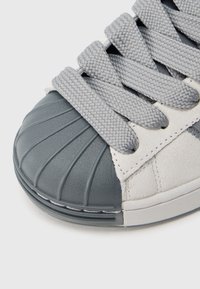 Tênis com biqueira de borracha cinza texturada com ranhuras, parte superior em tecido cinza e atacadores trançados cinza. Entressola branca lisa visível.