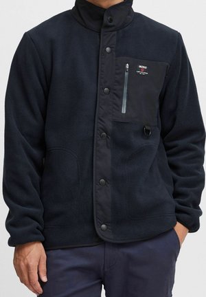 Man draagt een marineblauwe fleecejas met drukknopen, een borstzak met rits en label "INDIGO", en marineblauwe broek.