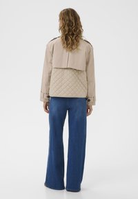 Kvinde med bølget hår, iført en beige quiltet jakke og blå brede jeans, stående med ryggen til mod en enkel baggrund.
