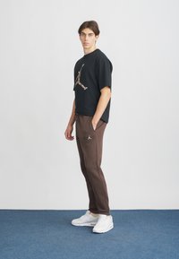 Schwarzes Baumwoll-T-Shirt mit Grafiklogo, braune Jogginghose mit Seitentaschen und Logo, kombiniert mit weißen Sportschuhen. Einfarbiger Hintergrund.