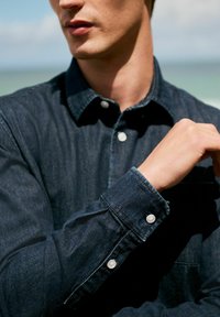 Donkerdenim overhemd met een knoopsluiting bij de kraag en manchetten, voorzien van witte knopen en een zakdetail, met een gladde textuur.