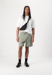 Vit oversized t-shirt, ljusgröna shorts, svart crossbody-väska, svarta sneakers med vita ränder och svarta strumpor. Enkel design.
