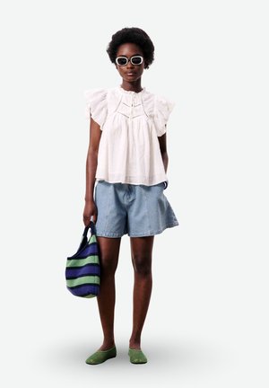 Vrouw met een witte blouse met ruches, lichtblauwe shorts, groene schoenen, witgekaderde zonnebril, houdend een marineblauw-groen gestreepte tas vast.