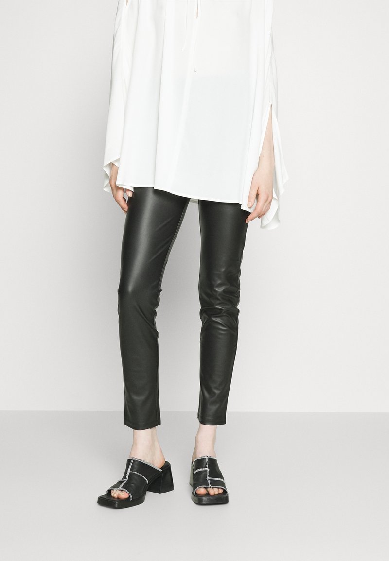 Femme portant un pantalon en cuir noir slim, des sandales noires à talons ouvertes à l’avant, et un haut blanc ample à manches longues avec des fentes sur les côtés.