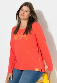 Langärmeliges Korallen-Shirt mit rundem Ausschnitt, gelben Grafikdesigns und Seitenschlitzen. Kombiniert mit blauen Jeans, auf einem gelben Hocker sitzend.