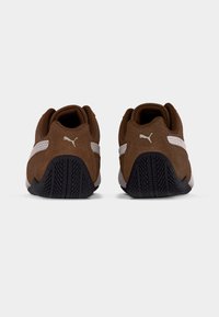 Brune ruskindssko med hvide detaljer og sorte gummisåler, med en tekstureret slidbane og et lavprofil-design.