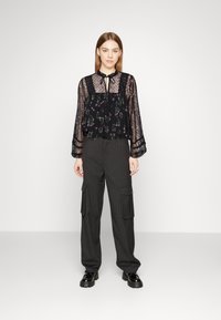 Blusa nera a fiori realizzata in tessuto trasparente con dettagli in pizzo, abbinata a pantaloni cargo a righe con vita alta. Scarpe nere lucide completano il look.
