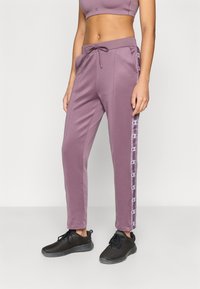 Μωβ αθλητικό παντελόνι με ελαστική μέση, κορδόνι και λογότυπο στα πλαϊνά. Λεπτό ύφασμα με εφαρμοστή γραμμή μέχρι τον αστράγαλο. Μαύρα αθλητικά παπούτσια.