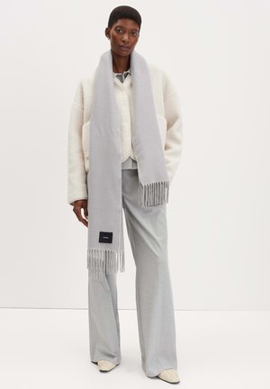 Écharpe grise avec des bords à franges, fabriquée en matériau doux, portée sur une veste crème et un pantalon gris clair. Comprend une petite étiquette de marque.