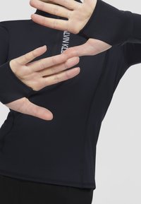 Osoba v černém dlouhém rukávu s otvory na palce, s rukama vztyčenýma vpřed, částečně si zakrývá tvář a horní část těla.