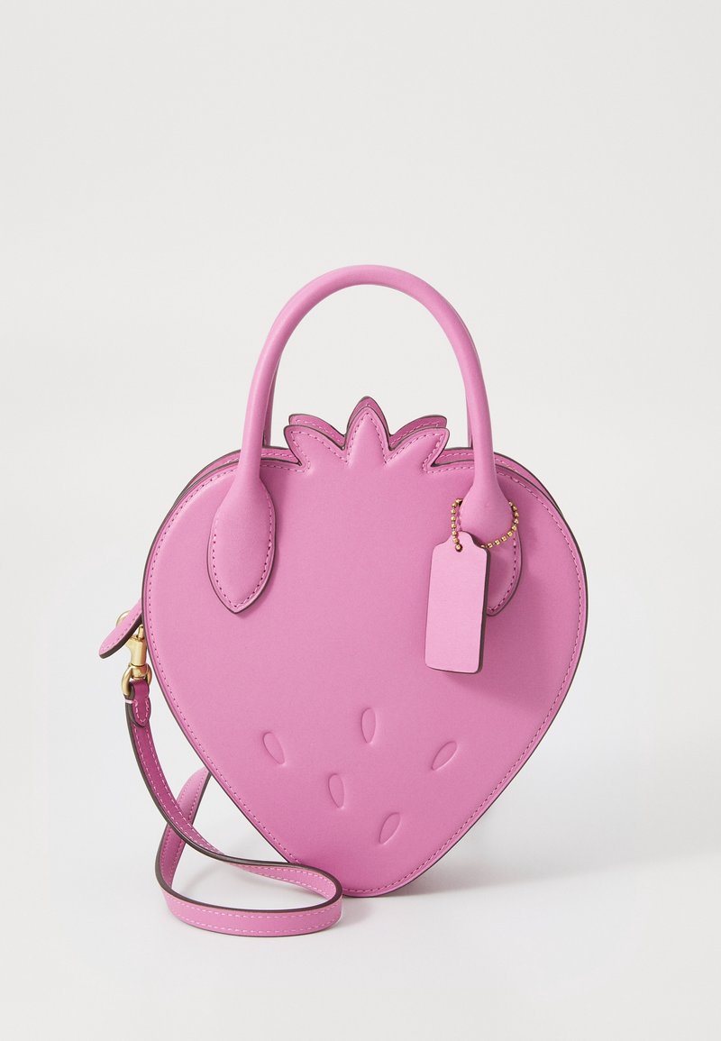 Sac à main en cuir rose en forme de fraise avec deux poignées, une bandoulière amovible et une petite étiquette suspendue.