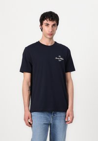 Marineblaue Baumwoll-T-Shirt mit kurzen Ärmeln, Rundhalsausschnitt und weißem gesticktem Logo auf der linken Brust. Kombiniert mit hellblauen Jeans.
