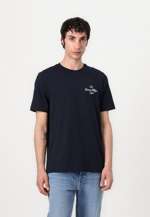 Navyblauwe katoenen T-shirt met korte mouwen, ronde hals en een wit geborduurd logo op de linkerkant van de borst. Gepaard met lichtblauwe spijkerbroek.