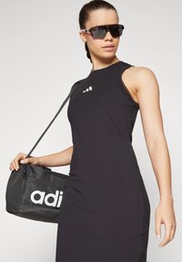 Vestito nero senza maniche con silhouette aderente, caratterizzato da tre strisce bianche sul petto. Borsa a secchiello nera con logo "adidas" bianco.
