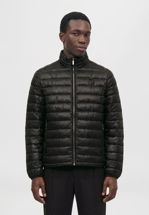Un homme porte une veste noire matelassée zippée jusqu’au cou, ornée d’un petit écusson sur la poitrine, sur un fond clair uni.