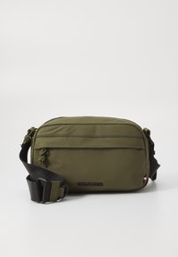 Olive green crossbody bag in durable fabric with a front zipper pocket, adjustable strap, and embossed logo. Features side accents.