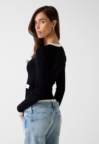 Zwart geribd sweater met een witte kraag en manchetten, voorzien van een kleine witte knoopdetail bij de taille, gecombineerd met lichtblauwe jeans.