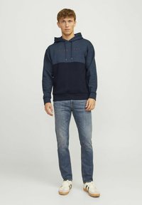 Sudadera con capucha en azul oscuro y marino, con un bolsillo frontal y cordones. Combinada con jeans de un azul claro y zapatillas beige.