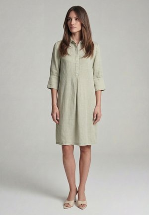 Frau steht in einem knielangen hellgrünen Leinenkleid mit Dreiviertelärmeln und beigen offenen Absatzschuhen vor einem einfarbigen Hintergrund.