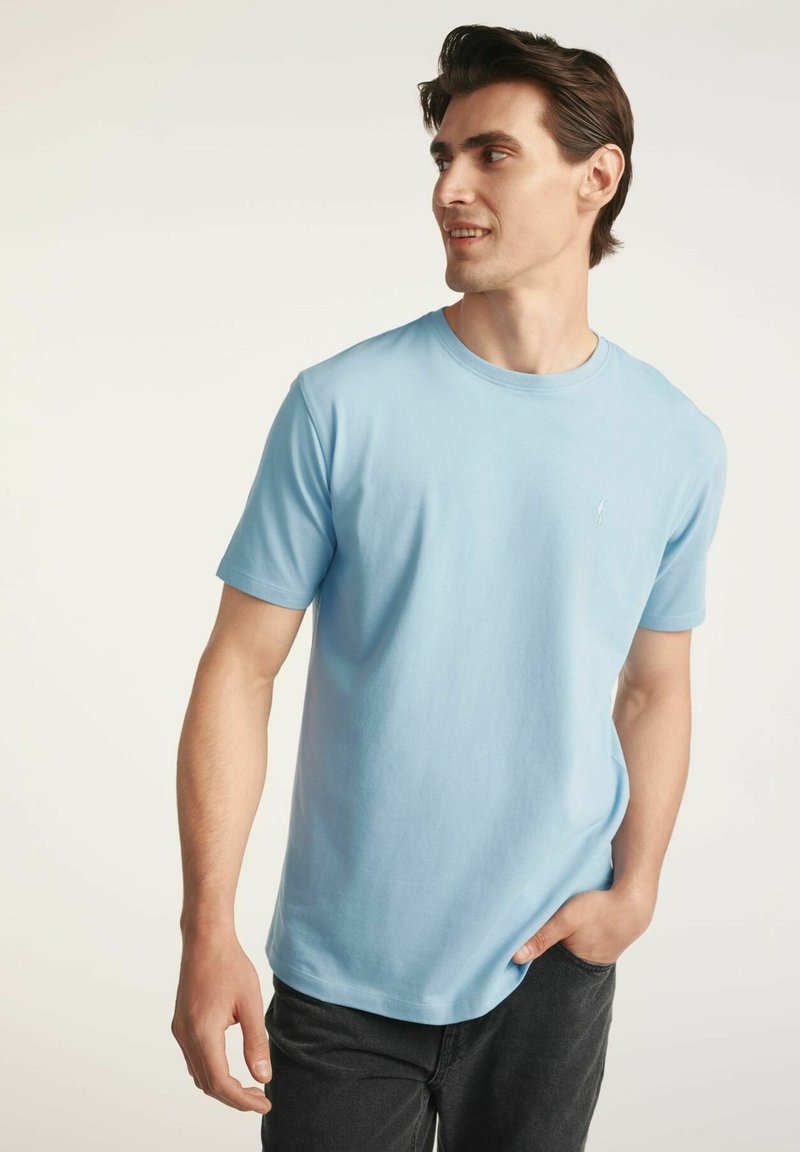 Jasnoniebieska bawełniana koszulka z krótkim rękawem i okrągłym dekoltem, z małym logo na piersi. Noszona z ciemnymi dżinsami.