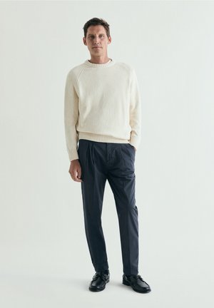 Hombre con un suéter de punto color crema, pantalones oscuros plisados y zapatos de cuero negro, de pie frente a un fondo claro y sencillo.
