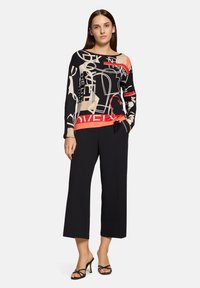Czarna bluzka z długim rękawem z abstrakcyjnym wzorem w beżu, czerwieni i czerni. Do tego czarne spodnie z szerokimi nogawkami oraz czarne sandały na paski.
