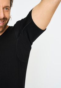 Zwart T-shirt met korte mouwen en een zachte textuur, met gebogen naden bij de oksels en een V-halsontwerp, gedragen door een persoon die een arm opsteekt.