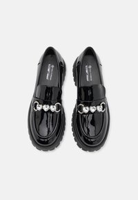 Mocassins en cuir verni noir avec une semelle épaisse, dotés d'une bride en chaîne ornée de strass en forme de cœur à l'avant.