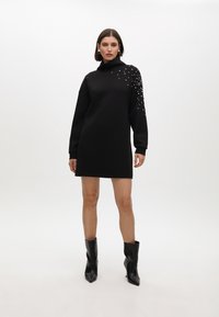 Schwarzes Sweatshirt-Kleid mit hohem Kragen, langen Ärmeln und mit Perlen an einer Schulter verziert, mit einem lockeren, entspannten Schnitt.