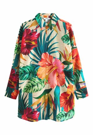 Kobieta z kręconymi włosami, ubrana w kolorową kwiecistą koszulę i koralowe figi bikini, stoi obok tropikalnych roślin, patrząc na bok.