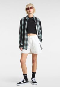 Kariertes Hemd in Schwarz- und Teal-Tönen, schwarzes kurzes Oberteil, weiße Shorts und schwarze Vans Sneakers mit weißen Schnürsenkeln und Logo-Socken.
