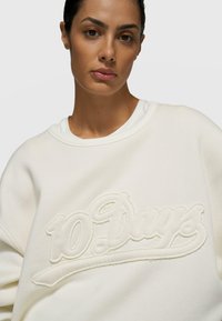 Sudadera blanca de gran tamaño con un bordado texturizado "10 Days" en color a juego, material de algodón suave y cuello redondo.
