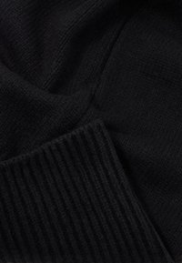 Černá pletená látka s texturovanou tkaninou a žebrovanými okraji. Materiál je měkký a mírně chlupatý, což zdůrazňuje útulný vzhled.