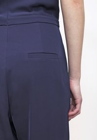 Námořnický modrý overal s hladkou texturou, s malou zadní kapsou a přiléhavým střihem. Tkanina vypadá lehce a elegantně.