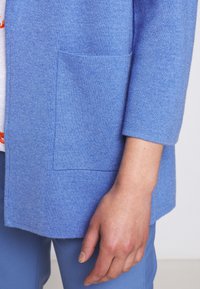 Blå texturerad kofta med ficka och trekvartsärmar, tillsammans med ljusblå byxor. Närbild av tygdetaljer och sömnad.